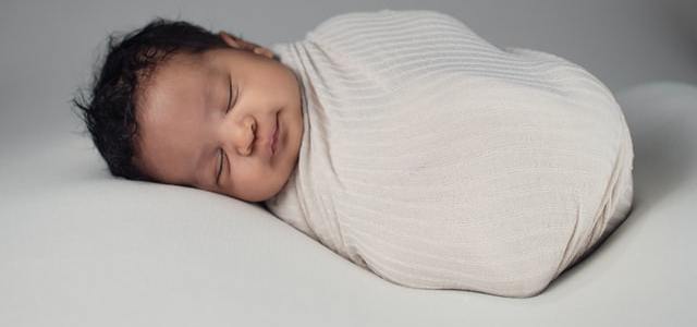 Baby with dark hair sleeping on white surface and wrapped in a white blanket.